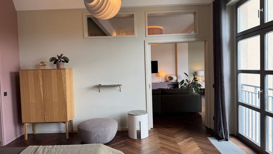 Bright modern apartment bedroom with herringbone wood floors, a large multi-pane window with dark curtains, and a sculptural pendant light. A wooden cabinet, round pouf, small side table, and a doorway with transom windows open into a living area beyond.