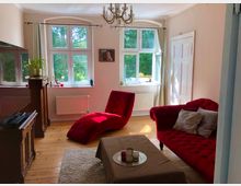 Wohnzimmer mit hellen Wänden und Holzboden. Große Fenster lassen Tageslicht herein, rote Polstermöbel stehen im Raum, und ein Kronleuchter hängt von der Decke. Ein Kamin und ein kleiner Tisch mit Dekoration vervollständigen die Einrichtung.