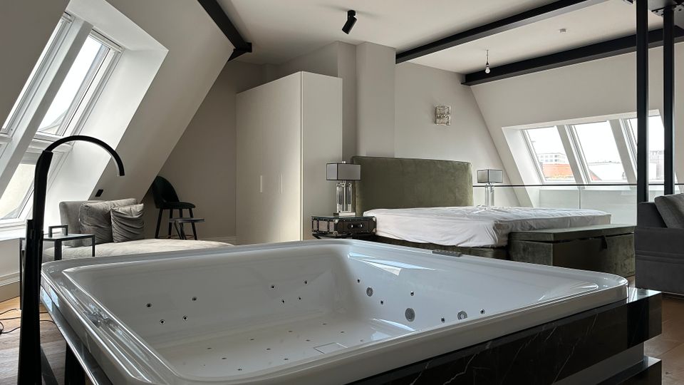 Modernes Dachgeschoss-Schlafzimmer mit freistehender Whirlpool-Badewanne im Vordergrund, schrägen Dachfenstern und sichtbaren schwarzen Deckenbalken. Im Hintergrund stehen ein großes Bett mit Nachttischen sowie ein Schrank; durch die Fenster ist ein Blick über die Stadt möglich.
