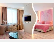 Ein modernes Hotelzimmer mit hellrosa und weißen Farbakzenten. Der Raum ist durch eine geschwungene, beleuchtete Trennwand mit eingebautem Flachbildfernseher in einen Arbeitsbereich mit Schreibtisch und einen Schlafbereich mit einem Doppelbett geteilt.