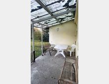 Überdachte Terrasse mit einem alten, teilweise durchsichtigen Dach aus Wellplatten, einem weißen Kunststofftisch, mehreren Stühlen aus Holz und Kunststoff sowie einer schlichten Wandlaterne. Im Hintergrund ist ein Garten mit Bäumen und Gras erkennbar.