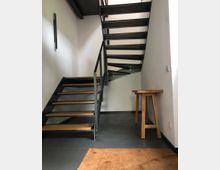Metalltreppe mit Holzstufen in einem modernen, schlicht gehaltenen Innenraum; links eine weiße Wand, rechts ein kleiner, hölzerner Beistelltisch und ein Fußboden aus dunklen Fliesen. Im Vordergrund liegt eine braune Fußmatte.