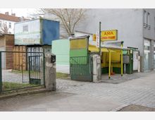 Im Bild ist ein einfacher Asia-Imbiss mit gelb-grüner Fassade und einem Schild mit der Aufschrift "Asia China Pfanne" zu sehen. Der Imbiss befindet sich in einem Hinterhofbereich mit Metallzaun und einem gepflasterten Boden.