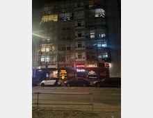 Mehrstöckiges Wohn- und Geschäftsgebäude bei Nacht, mit beleuchteten Balkonen in den oberen Etagen und Gewerbeeinheiten im Erdgeschoss, darunter ein Café, ein Sexshop und ein Sexkino mit leuchtenden Neon-Schildern. Vor dem Gebäude parken mehrere Autos entlang einer zweispurigen Straße.