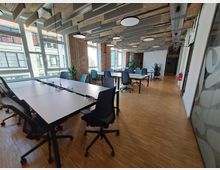 Ein moderner Büroraum mit mehreren Tischen und schwarzen Bürostühlen auf einem hellen Holzboden. Große Fenster lassen Tageslicht herein, Backsteinwände und freiliegende Deckenstrukturen verleihen einen industriellen Charakter; grüne Pflanzen lockern den Raum auf.