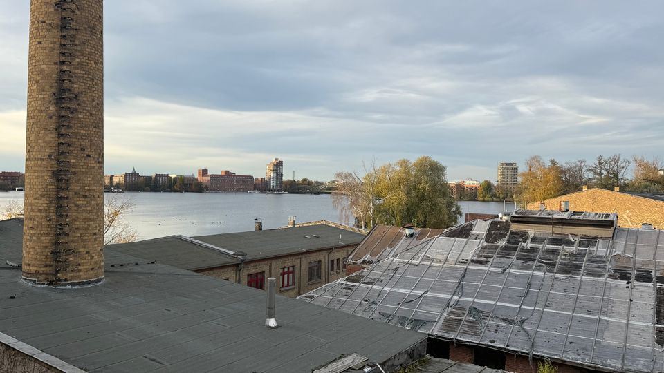 Blick über flache Industriedächer mit einem hohen gemauerten Schornstein im Vordergrund, daneben teils verwitterte Dachflächen mit Oberlichtern. Dahinter liegt ein breites Gewässer mit Uferbäumen und einer Stadt-Silhouette aus Wohn- und Gewerbebauten unter bewölktem Himmel.