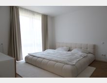 Schlafzimmer mit einem großen, gepolsterten Bett in hellem Beige, weißer Bettwäsche und zwei Kissen. Im Hintergrund befindet sich ein großes Fenster mit halbtransparenten Vorhängen, durch das Tageslicht hereinfällt.