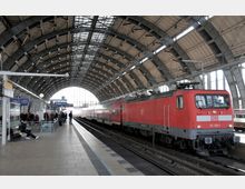 Ein moderner Bahnhof mit einer großen, gewölbten Glas- und Stahlkonstruktion; ein roter Regionalzug der Deutschen Bahn steht am Bahnsteig, während Passagiere warten oder sich bewegen. Im Hintergrund ist ein Uhrenturm und weitere Bahnhofsbeschilderung zu sehen.