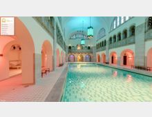 Hallenbad mit einem rechteckigen Schwimmbecken, umgeben von weißen Bögen und hohen Wänden im klassischen Stil. Oberhalb der Bögen befinden sich verziertes Steinwerk und große Fenster, während grün beleuchtete Hängeleuchten die Decke schmücken.