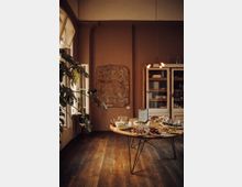 Ein gemütlicher Innenraum mit Holzboden und warmen, braunen Wänden. Im Vordergrund steht ein runder Holztisch mit verschiedenen Schalen und Tellern voller Fingerfood, während im Hintergrund ein Kunstwerk an der Wand und ein Schrank mit Geschirr zu sehen sind.