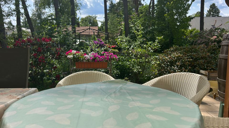 Terrasse oder Garten-Sitzplatz mit rundem Tisch (hellgrüne Tischdecke) und Korbstühlen, umgeben von dichtem Grün und blühenden Sträuchern. Im Hintergrund stehen hohe Bäume, ein Metall-Pavillonrahmen und Dächer von Wohnhäusern unter blauem Himmel.
