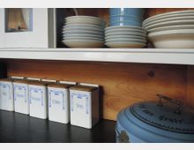 Ein Küchenschrank mit Holzhintergrund zeigt im oberen Regal gestapelte Teller und Schüsseln, teils in Weiß und Blau. Im unteren Bereich stehen beschriftete weiße Vorratsdosen mit blauen Verzierungen, daneben eine runde, blaue Backform mit Deckel.