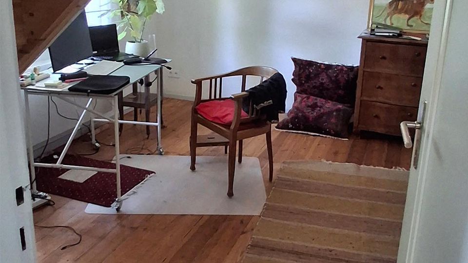 Blick in ein kleines Dachgeschosszimmer mit schräger Holzdecke und Holzdielenboden. Links steht ein einfacher Schreibtisch mit Monitor und Pflanze am Fenster, rechts eine Kommode mit Bild sowie Sitzkissen und ein Stuhl auf Teppichen.