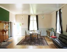 Ein heller Wohnraum mit Holzboden, einem rechteckigen Teppich, einem Esstisch und vier Stühlen in der Mitte. Links steht eine Garderobe aus Holz, rechts ein schwarzes Ledersofa und eine Kommode mit Keramiktöpfen; große Fenster mit Vorhängen lassen Tageslicht herein.