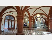 Innenansicht einer großen Halle mit roten Backsteinsäulen und kreuzrippenartigen Rundbögen, die eine helle, gewölbte Decke tragen. Der Raum hat ein gemustertes Fliesenbodenmosaik, hängende Kugelleuchten sowie Glastüren und Durchgänge zu angrenzenden Bereichen.