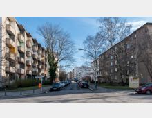 Wohnstraße in einem städtischen Gebiet mit mehrstöckigen Wohngebäuden auf beiden Seiten, vereinzelt Bäumen und parkenden Autos entlang des Straßenrandes. Im Hintergrund befinden sich weitere mehrstöckige Gebäude, und der Himmel ist klar und blau.