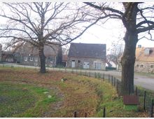 Ländliche Szene mit mehreren alten Stein- und Ziegelgebäuden im Hintergrund, umgeben von kahlen Bäumen. Im Vordergrund ein eingezäuntes, grasbewachsenes Gelände mit Laubblättern auf dem Boden.
