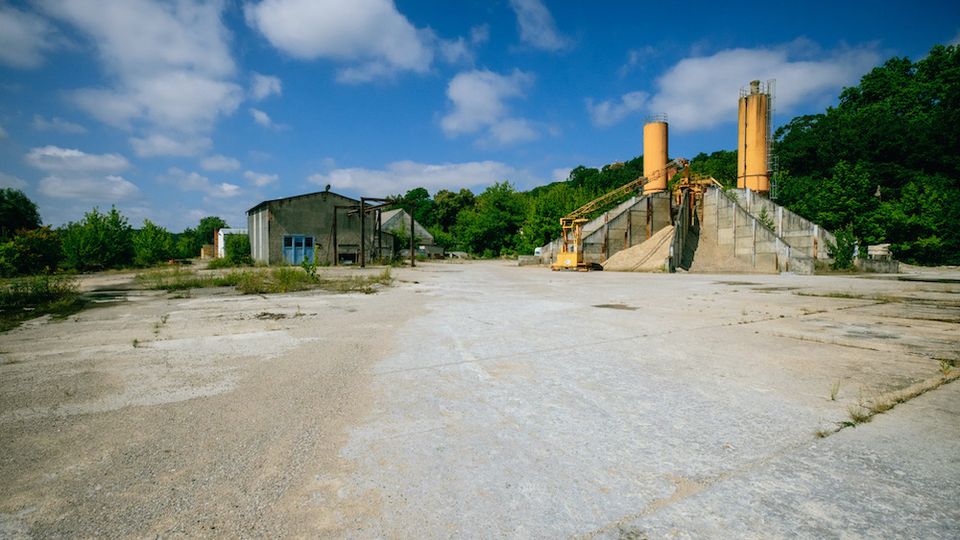 Weitläufiges Gelände mit Betonboden, einer verlassenen Industrieanlage mit rostfarbenen Silotürmen rechts und einem kleinen, verfallenen Gebäude links, umgeben von vereinzelter Vegetation und Wald im Hintergrund unter blauem Himmel.