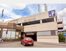 Mehrstöckiges Parkhaus mit einem beschilderten Eingang zur Tiefgarage „Q-Park Märkische Zeile“, daneben ein Geschäftseingang mit moderner Glasfassade. Im Vordergrund fährt ein rotes Auto, im Hintergrund sind Logos von Kaufland und Aldi sichtbar.