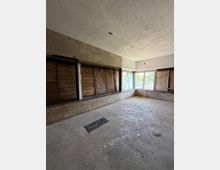 Ein leerer Raum mit unverputzten Wänden und Betonboden, großen Fenstern an einer Ecke und sichtbaren Holzvertäfelungen entlang der unteren Wandbereiche. Die Umgebung wirkt unfertig oder renovierungsbedürftig.