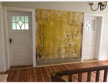 Ein Innenraum mit Holzdielenboden, einer gemusterten Wandmalerei mit gelbem Hintergrund und figürlichen Darstellungen in der Mitte, flankiert von zwei weißen Türen mit Glaseinsätzen. Im Vordergrund ein hölzernes Treppengeländer und an der Decke eine schlichte Hängelampe.