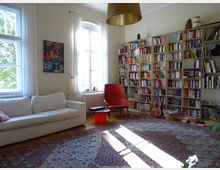 Helles Wohnzimmer mit einer weißen Couch, einem roten Sessel und einem großen Bücherregal, das die rechte Wand füllt. Zwei große Fenster mit weißen Vorhängen lassen reichlich Tageslicht herein, und auf dem Boden liegt ein großer gemusterter Teppich.