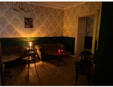 Gemütliches Café oder Lounge mit gedämpfter Beleuchtung, Holzboden und Vintage-Ambiente. Der Raum ist mit dunklen Chesterfield-Sofas, kleinen Holztischen, einem Kronleuchter und floraler Tapete dekoriert.