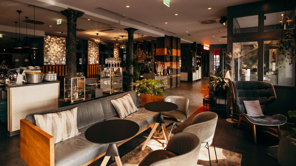 Moderner Lounge- und Cafébereich mit Sofas, gepolsterten Sesseln und runden Beistelltischen auf einem Teppich. Im Hintergrund stehen eine Bar-/Kaffeetheke mit Glasvitrinen, Regale, schwarze Säulen, Pflanzen und warme Deckenbeleuchtung in einem offenen Innenraum.