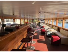 Innenraum eines Restaurantschiffs mit Holzausstattung, einer Bar auf der linken Seite und Sitzbereichen mit Tischen und gepolsterten Bänken auf beiden Seiten. Große Fenster und Oberlichter bieten einen Ausblick auf das umliegende Wasser.