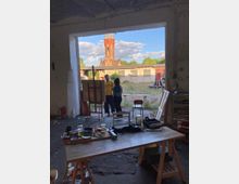 Innenraum eines Ateliers mit einem Holztisch voller Malutensilien im Vordergrund. Im Hintergrund ist ein offener Torbogen, durch den eine Backsteinarchitektur mit Türmen und eine Hofanlage zu sehen sind. Zwei Personen stehen im Tor und unterhalten sich.