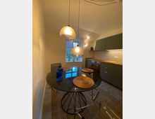 Moderne Küche mit einem Essbereich, bestehend aus einem runden, schwarzen Glastisch mit geflochtenen Tischsets und transparenten Stühlen. Die Küche verfügt über dunkelgraue Schränke, eine Holzarbeitsplatte, Hängelampen und ein Fenster mit Blick ins Freie.