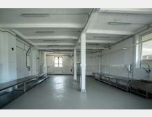 Weiß gefliester, leerer Industriebereich mit hohen Decken und sichtbarer Verkabelung; ausgestattet mit Edelstahl-Arbeitsflächen und Waschbecken entlang der Wände. Große Fenster lassen Tageslicht herein, und die Umgebung wirkt funktional und sauber.