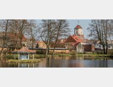 Ein kleiner Teich mit einer überdachten Holzplattform auf einer Insel im Vordergrund, umgeben von kahlen Bäumen. Im Hintergrund sieht man eine Kirche mit rotem Kuppeldach sowie umliegende Gebäude mit roten und grauen Ziegeldächern.