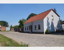 Einfamilienhaus mit weiß verputzter Fassade und rotem Satteldach, gelegen an einer gepflasterten Straße in einer ländlichen Umgebung. Im Hintergrund sind Bäume, ein Zaun und kleine Holzbauten zu sehen.