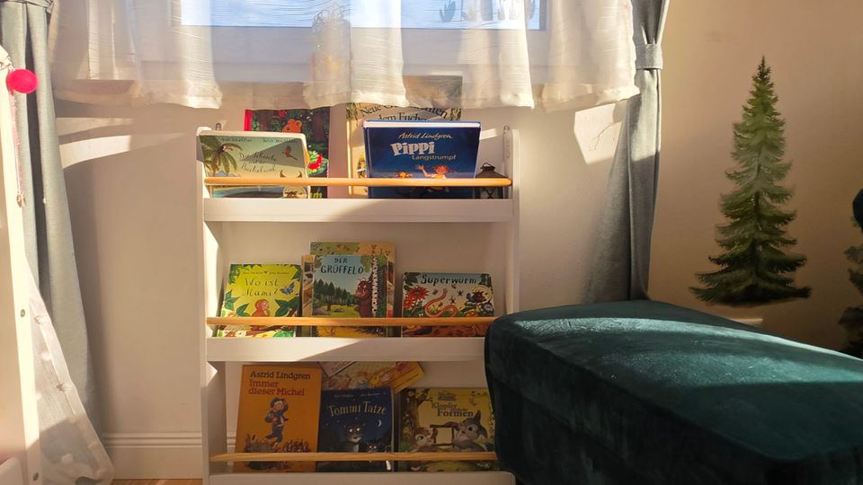 Helle Leseecke in einem Kinderzimmer mit großem Fenster, weißen Gardinen und grauen Vorhängen sowie Schmetterlingsaufklebern am Glas. Darunter steht ein niedriges, weißes Bücherregal mit Bilderbüchern; davor liegt ein heller Teppich auf Holzboden, daneben ein dunkelgrüner Sitzhocker und eine Wanddeko in Form eines kleinen Baums.