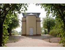 Gelb gestrichener, kleiner Pavillon mit einer weißen Tür und einem gestreiften Dach, umgeben von Bäumen und einem sandigen Weg, der durch eine Laubenöffnung in den Vordergrund führt.