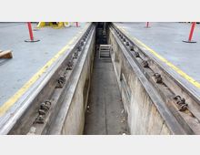 Eine Werkstattgrube mit Schienen und gelb markierten Sicherheitskanten, umgeben von einer Betonfläche. Im Hintergrund befindet sich eine Leiter innerhalb der Grube sowie Baustellenausrüstung und Absperrpfosten.