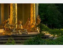 Goldene Skulpturen auf einer verzierten, mit Blattgold bedeckten Säule vor einem historischen Gebäude, umgeben von grüner Vegetation. Die Figuren stellen in orientalischem Stil gekleidete Menschen dar und sitzen auf einem steinernen Sockel, der von einer Treppe umgeben ist.