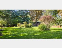 Großer Garten mit einer gepflegten Rasenfläche, umgeben von dichten Bäumen und Sträuchern. Im Hintergrund stehen eine Holzbank, ein Holzunterstand und eine Schaukel, während dekorative Pflanzen und Blumentöpfe den Vordergrund ergänzen.