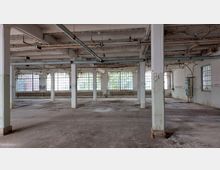 Industriehalle mit einer weitläufigen, leeren Fläche, weißen Säulen und sichtbaren Rohrleitungen an der Decke. Die großen Fenster an den Wänden lassen viel Licht einfallen, während die Wände und Böden deutliche Gebrauchsspuren zeigen.