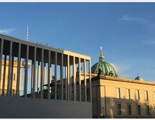 Moderne Architektur mit Säulen im Vordergrund, dahinter eine historische Kirche mit grünlicher Kuppel und goldener Spitze. Im Hintergrund ist ein Teil des Berliner Fernsehturms sichtbar, unter einem klaren blauen Himmel.