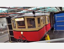Roter und beiger Zugwagen eines historischen S-Bahn-Zugs in einer Halle mit blauem Hintergrund und Stahlträgern an der Decke. Im Frontfenster ist die Beschilderung "Ring über Stralau-Rummelsburg" zu sehen.