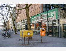 Straßenszene mit einem modernen Gebäude mit großen Glasfronten im Hintergrund, gelben Briefkästen und einem orangefarbenen Mülleimer im Vordergrund. Daneben befinden sich Fahrradständer mit geparkten Fahrrädern und Straßenschilder für die "Deitmerstraße" und "Schloßstraße".