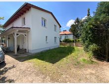 Zweistöckiges Wohnhaus mit weißer Fassade und rotem Dach, ergänzt durch einen kleinen überdachten Eingangsbereich. Das Haus steht auf einem Grundstück mit einem unbefestigten Zufahrtsweg, angrenzendem Rasen und Bäumen im Hintergrund.