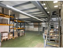 Lagerhalle mit hohen Regalen, auf denen Paletten mit verpackter Ware gestapelt sind. Im Vordergrund eine Metalltreppe, die zu einer Zwischendecke aus Stahl führt, und eine grüne Bodenfläche.