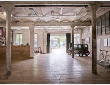 Großer, offener Innenraum einer ehemaligen Industriehalle mit Holzfußboden, freiliegenden Backsteinwänden und Säulen. Ein Oldtimer und ein Fahrrad stehen nahe der rechten Wand; eine große Glastür mit Blick auf einen Außenhof befindet sich im Hintergrund.