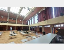 Großer Veranstaltungssaal mit Holzboden, bestückt mit weißen Tischen und schwarzen Stühlen. Der Raum hat eine hohe Decke mit großen Fenstern, roten Vorhängen, modernen Beleuchtungselementen sowie Sichtschutz und eine Bühne im Vordergrund mit Flaggen in der Ecke.