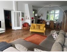 Wohnzimmer mit Parkettboden und moderner Einrichtung, darunter ein graues Ecksofa, ein gelbes Sofa und ein roter Teppich. Im Hintergrund ein Esstisch mit Stühlen, große Fenster mit Blick ins Grüne und eine übergroße Bogenlampe.