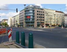 Straßenkreuzung in einer Stadt mit mehrstöckigen Gebäuden im Hintergrund, darunter moderne und ältere Fassaden. Im Vordergrund befinden sich Baustellenbereiche mit Absperrungen und Pollern, während Straßenbahndrähte und Verkehrsampeln die Szene überqueren.