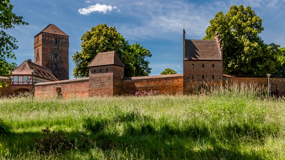 Blick über eine Wiese auf eine mittelalterliche Backsteinburg mit Ringmauer und mehreren Türmen, darunter ein hoher quadratischer Hauptturm. Im Hintergrund stehen große Laubbäume unter blauem Himmel, links ist ein Fachwerkgebäude an die Anlage angebaut.